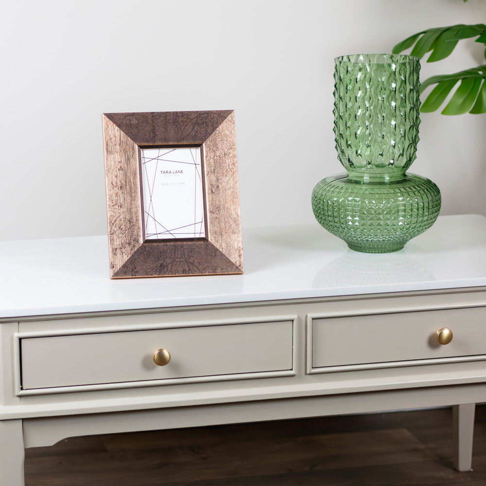 A square picture frame with a textured, metallic rose-gold/copper-brown finish sits on Monroe Coffee Table in Stone console table next to a green glass vase. The background is a clean white wall with a leaf visible in the upper right corner.