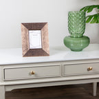 A square picture frame with a textured, metallic rose-gold/copper-brown finish sits on Monroe Coffee Table in Stone console table next to a green glass vase. The background is a clean white wall with a leaf visible in the upper right corner.