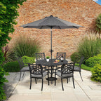 A 7-piece outdoor patio dining set with a round black table, four matching chairs, and a large black patio umbrella, arranged on a stone patio in front of a brick wall.