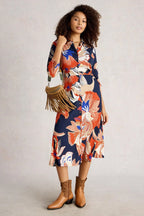 Woman wearing a colorful floral dress with brown boots and a fringed bag on a plain background