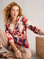 Woman wearing a colorful patterned shirt sitting on a wooden stool.