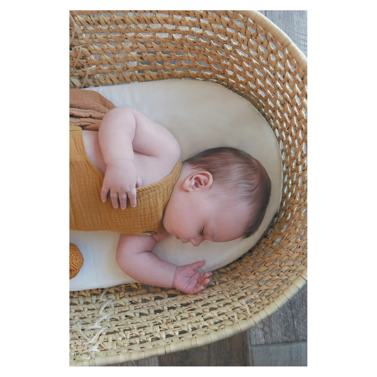 A sleeping baby wearing a mustard yellow knit romper and matching hat, resting inside a woven rattan bassinet on a cream cushion.
