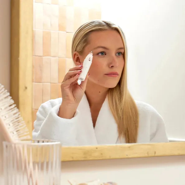 Woman in a white robe using a skincare device in front of a mirror.