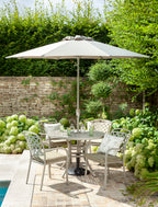 An outdoor patio scene with a light beige patio umbrella over a round glass-top dining table and four matching metal chairs with cushions. The setting includes a stone wall, lush green plants, and a tiled patio surface.