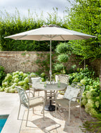 Outdoor patio dining set with a round metal table, four light gray metal chairs with cushions, a large beige patio umbrella, and a stone wall with greenery in the background.
