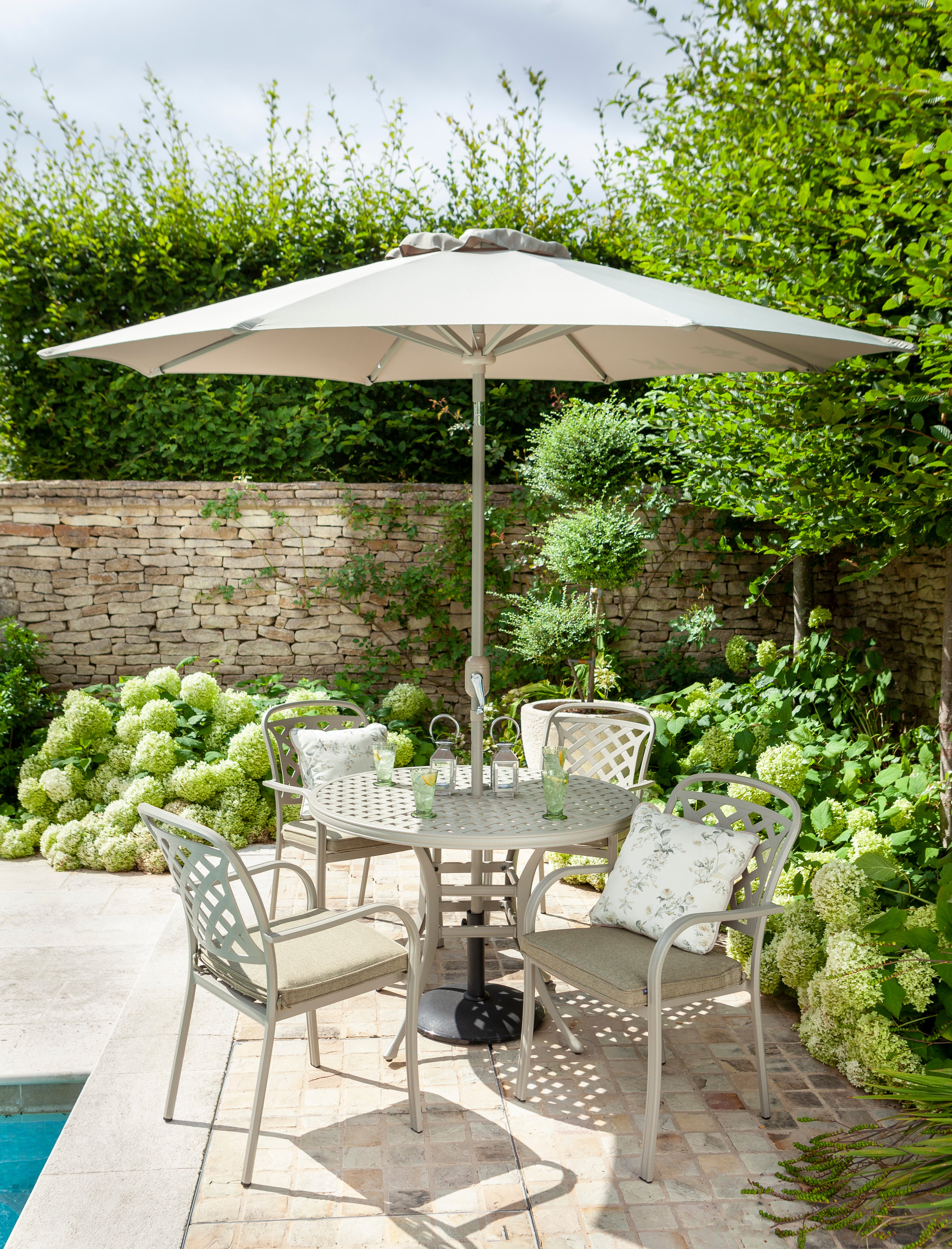 Outdoor patio dining set with a round metal table, four light gray metal chairs with cushions, a large beige patio umbrella, and a stone wall with greenery in the background.