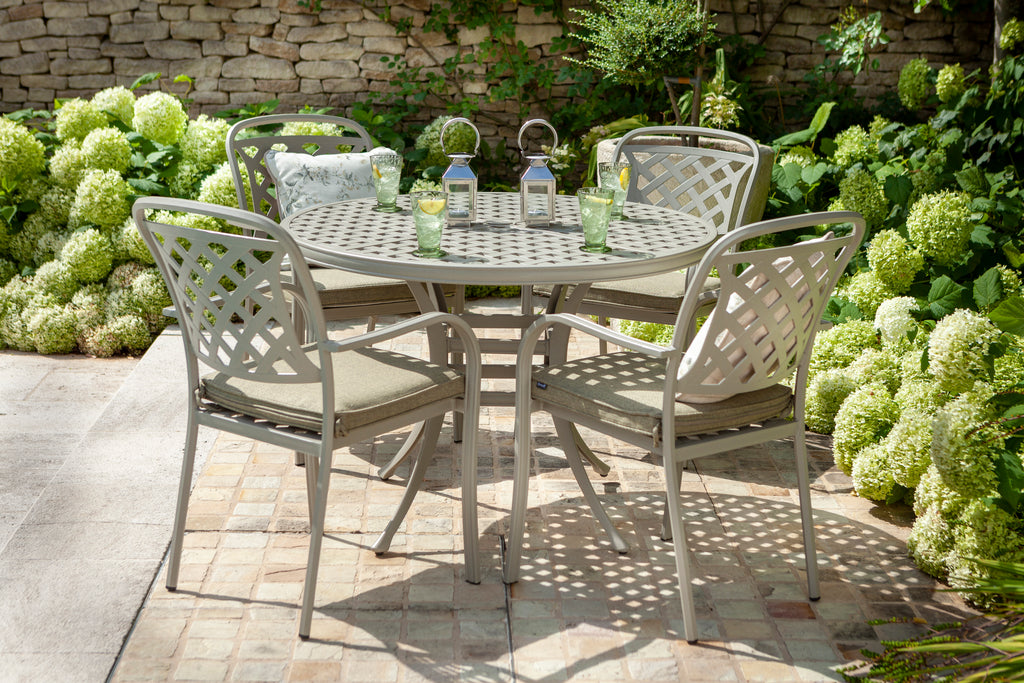 A 5-piece outdoor patio dining set featuring a round metal table and four matching lattice-back chairs with maize wheat  cushions, set on a stone patio surrounded by green hydrangea bushes. 