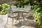 A 5-piece outdoor patio dining set featuring a round metal table and four matching lattice-back chairs with maize wheat  cushions, set on a stone patio surrounded by green hydrangea bushes. 