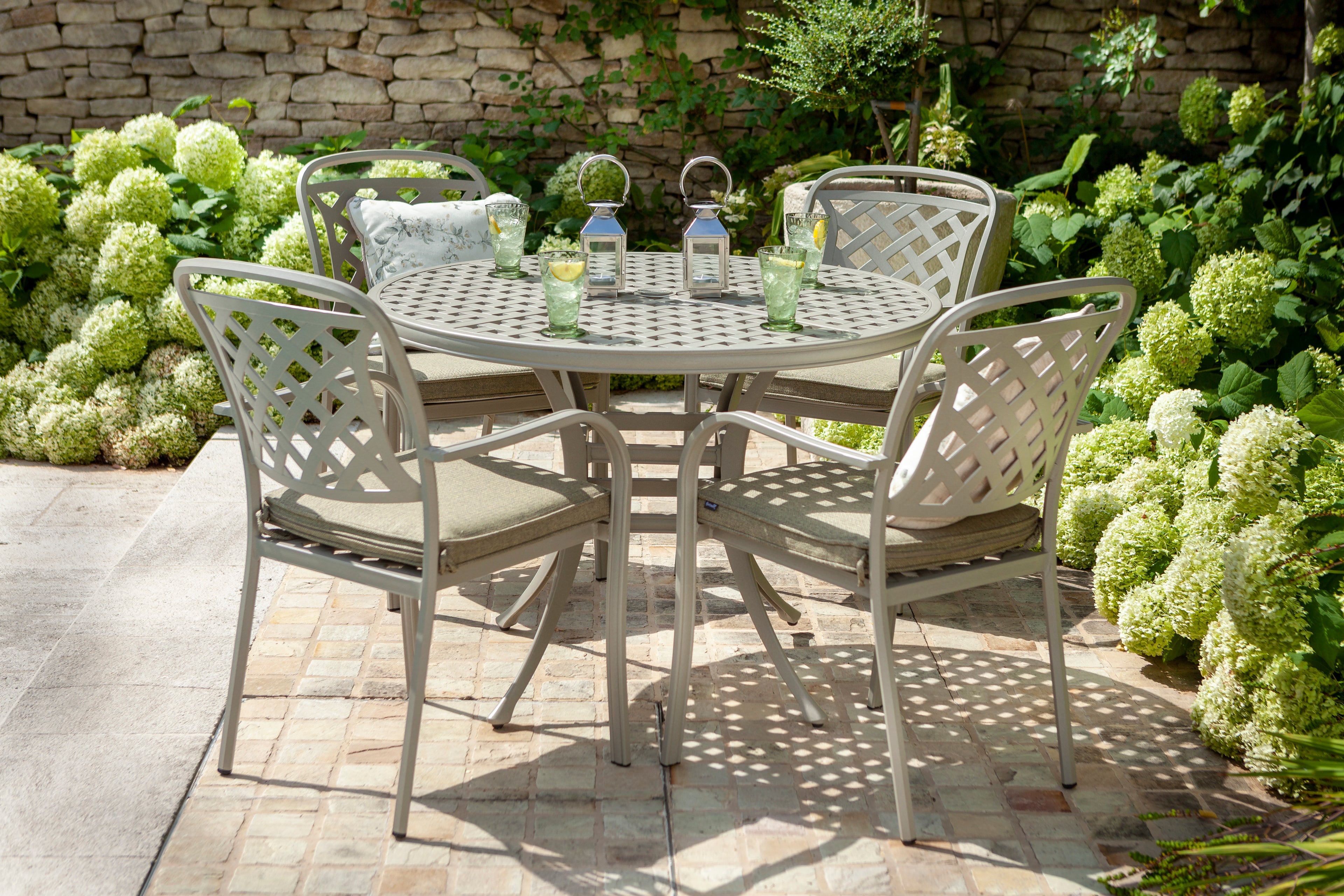A 5-piece outdoor patio dining set featuring a round metal table and four matching lattice-back chairs with maize wheat  cushions, set on a stone patio surrounded by green hydrangea bushes. 