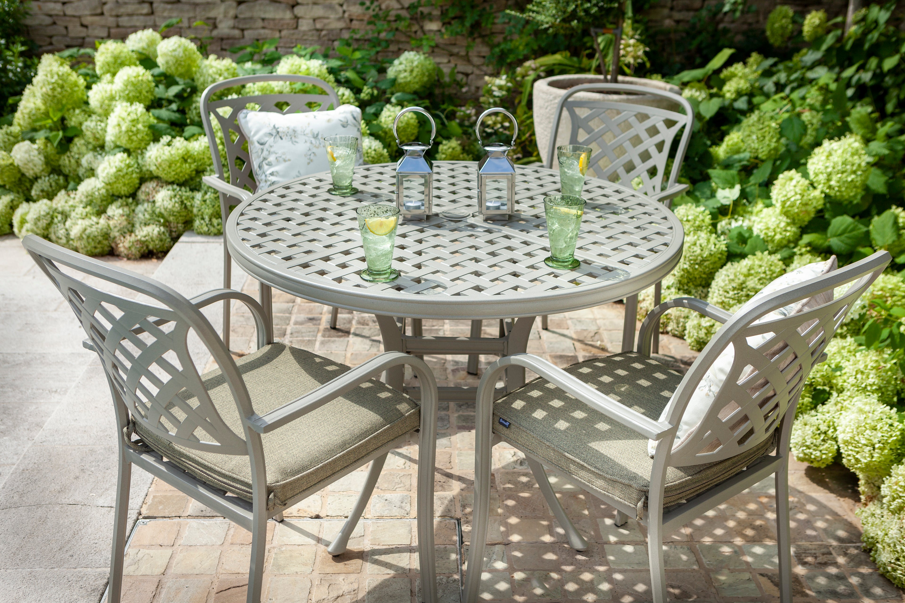 A 5-piece outdoor patio dining set featuring a round metal table and four matching chairs with cushioned seats, styled on a stone patio with green hydrangeas in the background.