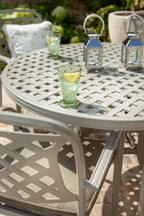 A round outdoor patio dining set with a white lattice-pattern metal table and matching chairs, styled with two clear drinking glasses containing green tea and lemon slices, and two silver lanterns on a stone patio.