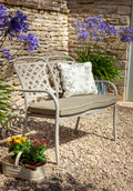 A light gray metal patio bistro chair with a lattice backrest and a maize wheat cushion, styled on a gravel patio with a patterned throw pillow, stone wall, and purple flowers.