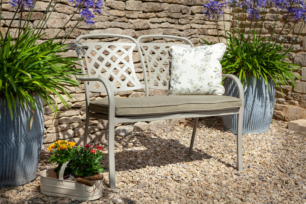 A white metal garden bench with two lattice-back seats and a cushioned seat and backrest, styled in a stone patio setting with potted plants and a decorative pillow.