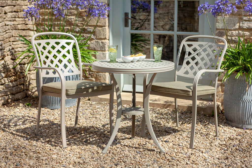 A three-piece outdoor patio bistro set featuring a round metal table and two matching armchairs with lattice backs and cushioned seats, styled on a gravel patio in front of a stone wall and glass door.