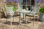 A three-piece outdoor patio bistro set featuring a round metal table and two matching armchairs with lattice backs and cushioned seats, styled on a gravel patio in front of a stone wall and glass door.