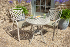 A 3-piece outdoor patio dining set featuring a round metal table and two matching armchairs with lattice backs, set on a gravel patio in front of a stone wall.