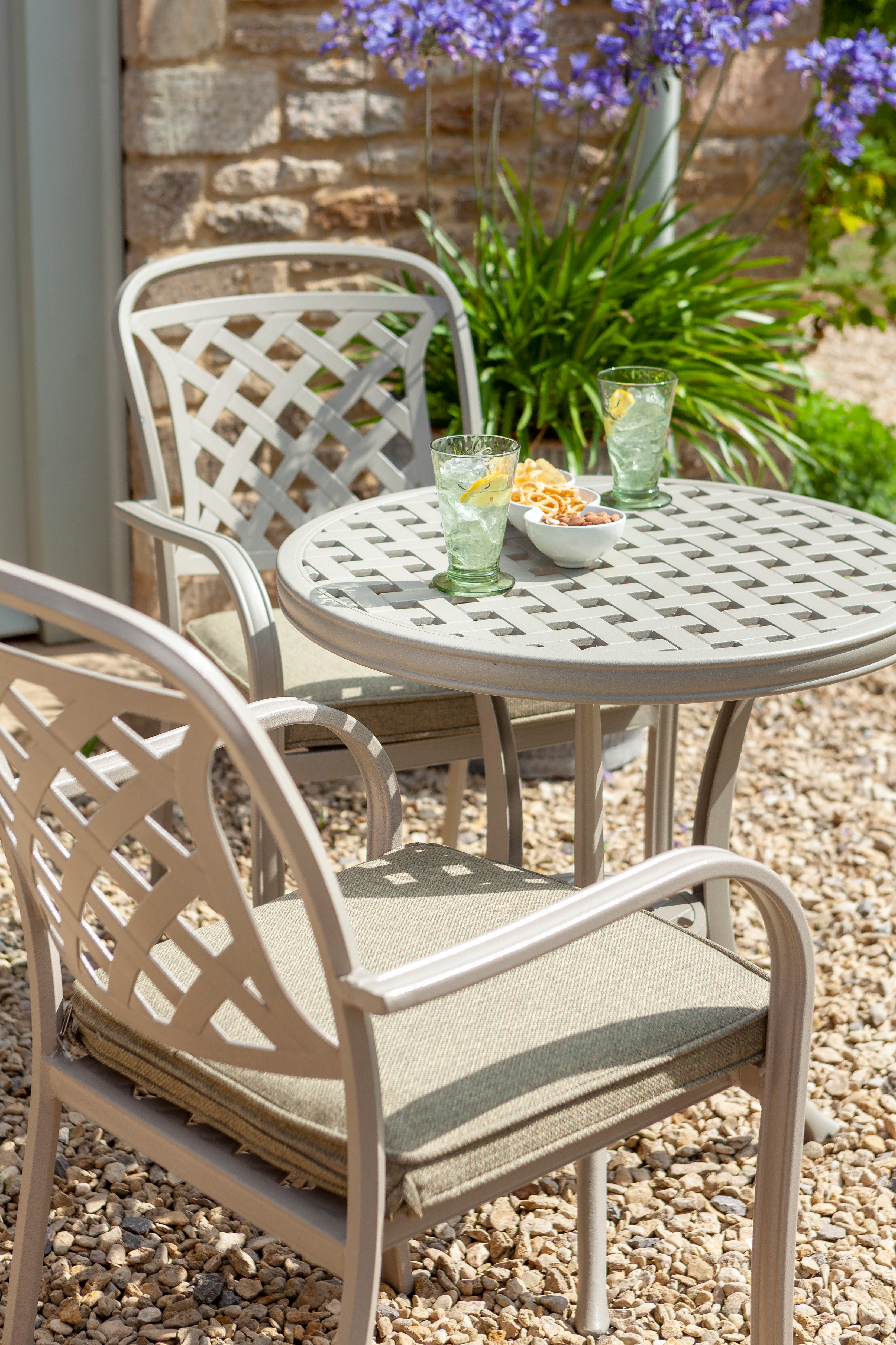 A light beige metal outdoor bistro set with a round lattice-patterned table and two matching armchairs with cushioned seats, shown on a gravel patio with green plants and purple flowers in the background.