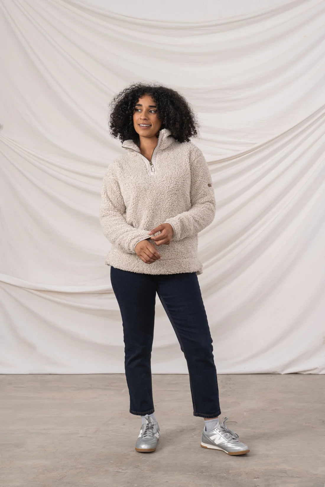 Woman wearing a beige sweater and dark pants standing against a plain white background