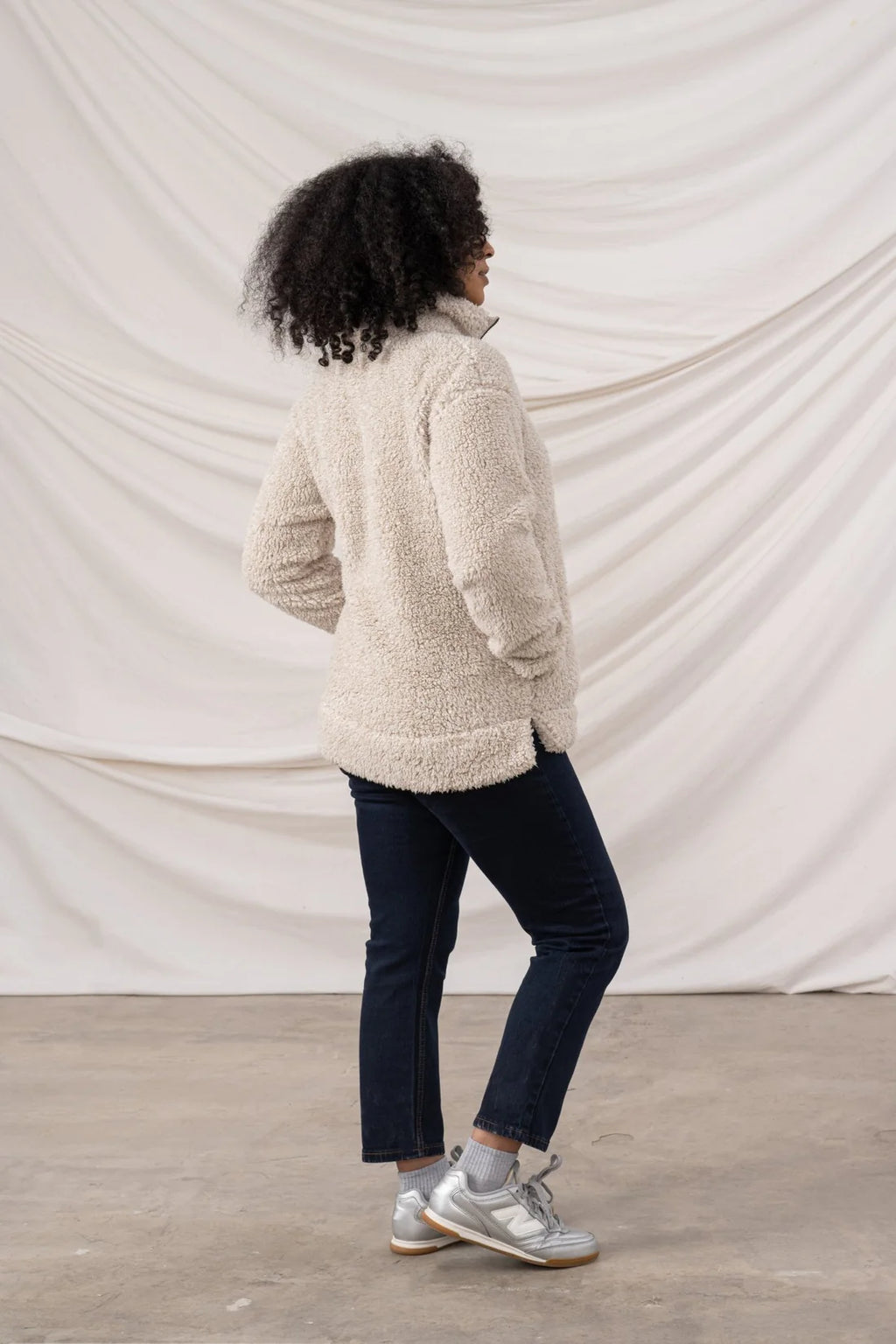 Person wearing a beige fleece jacket and dark jeans standing against a plain white background