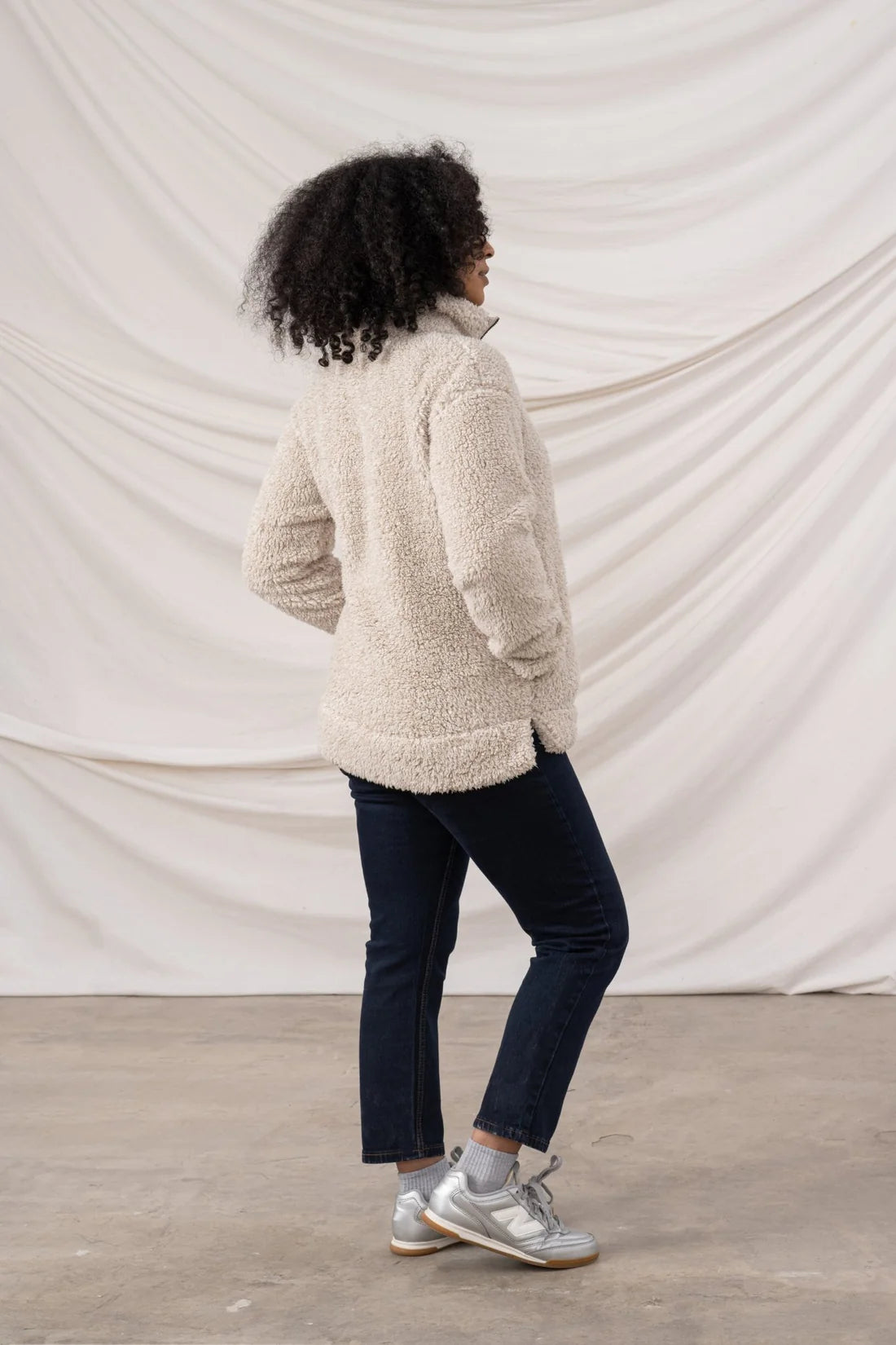 Person wearing a beige fleece jacket and dark jeans standing against a plain white background