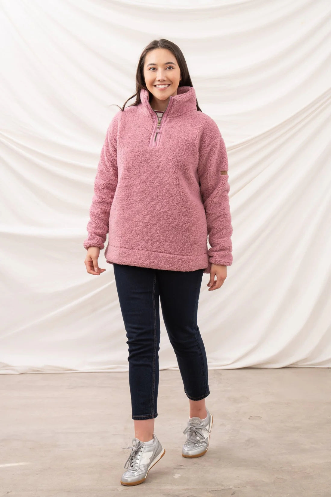 Woman wearing a pink pullover with a zipper, dark pants, and silver sneakers against a white backdrop.