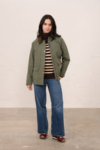 Woman wearing a green jacket, striped shirt, and blue jeans against a plain background