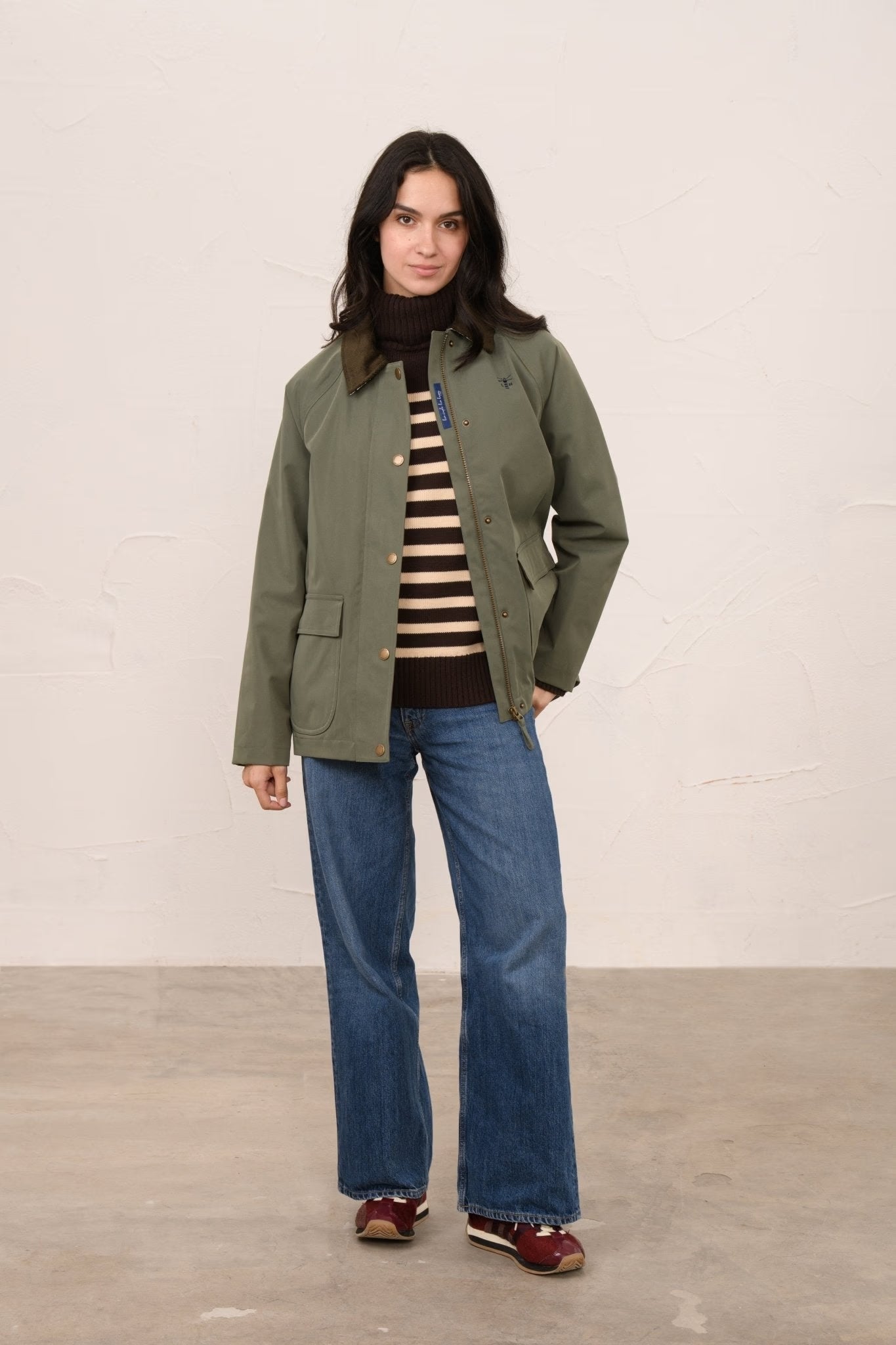 Woman wearing a green jacket, striped shirt, and blue jeans against a plain background