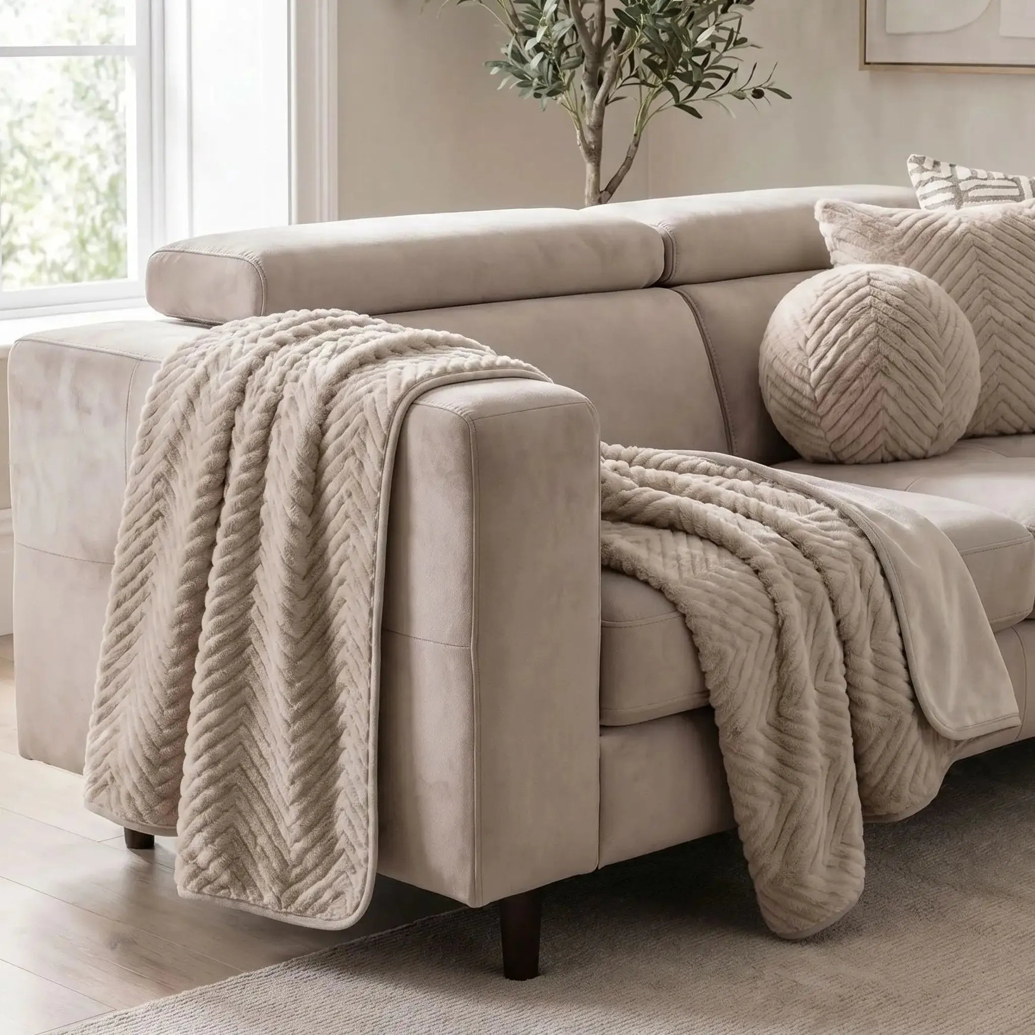 A light beige sofa in a bright living room, styled with a chunky cEllis Throw in Minkdraped over the arm and seat, and a matching round throw pillow.