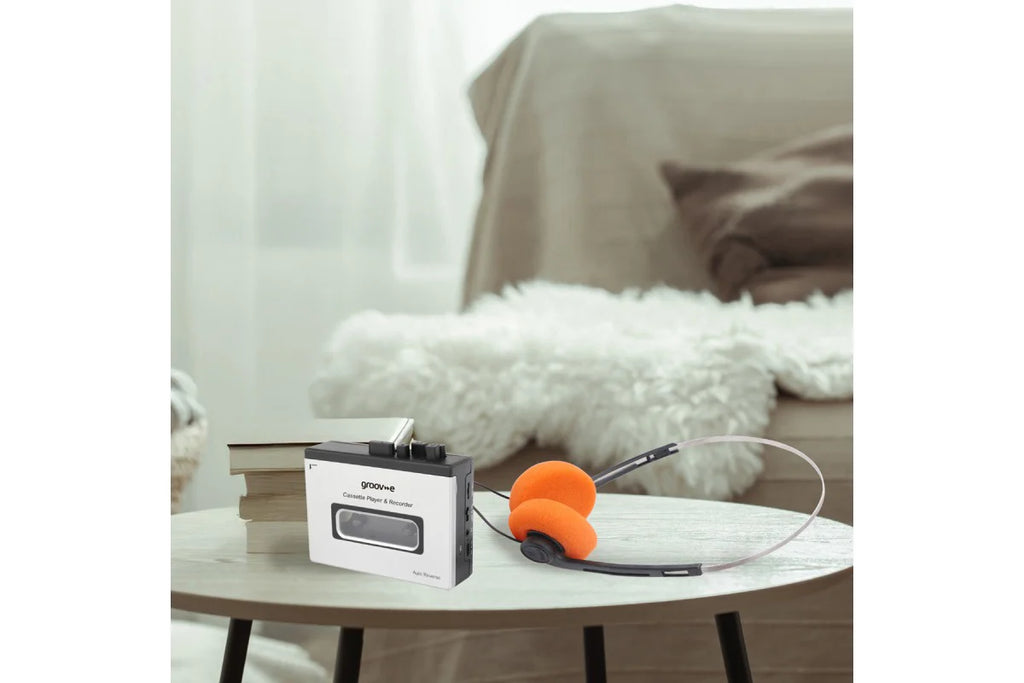 A portable cassette-style audio player with a white and black body sits on a round table in a softly lit living room. An orange over-ear headset is placed beside it, with a sofa and fluffy white throw in the background.