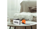 A portable cassette-style audio player with a white and black body sits on a round table in a softly lit living room. An orange over-ear headset is placed beside it, with a sofa and fluffy white throw in the background.