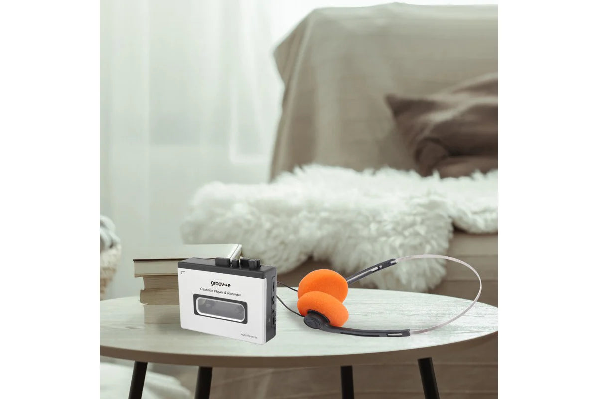 A portable cassette-style audio player with a white and black body sits on a round table in a softly lit living room. An orange over-ear headset is placed beside it, with a sofa and fluffy white throw in the background.