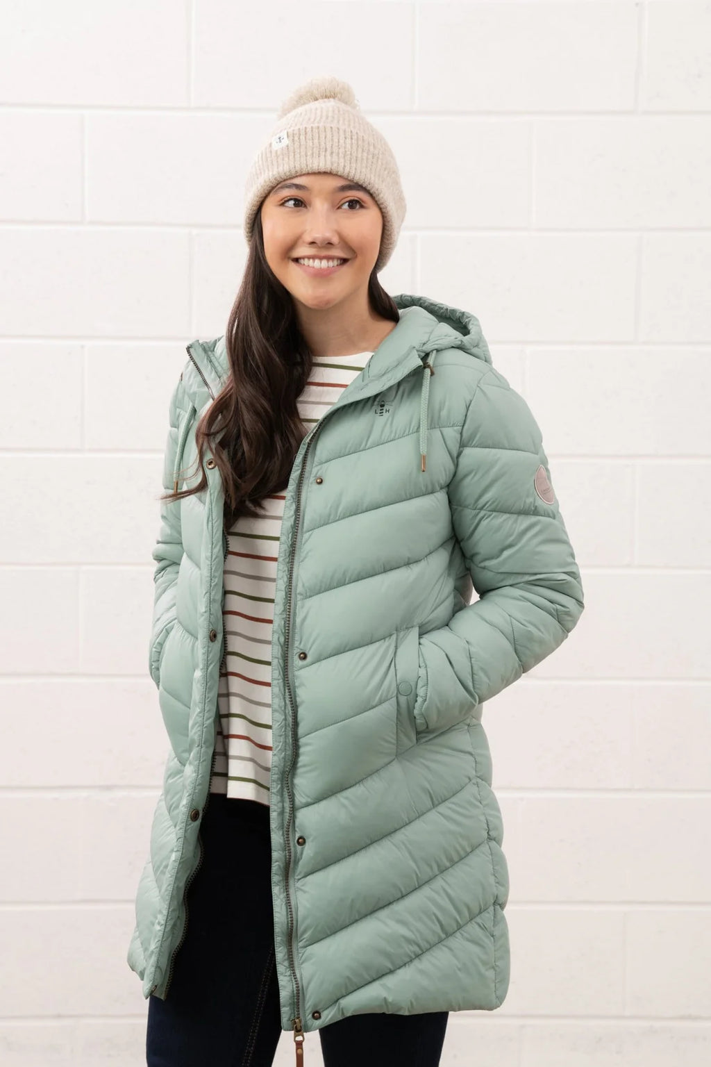 Woman wearing a long green puffer coat and a beanie against a white brick wall.