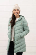 Woman wearing a long green puffer coat and a beanie against a white brick wall.