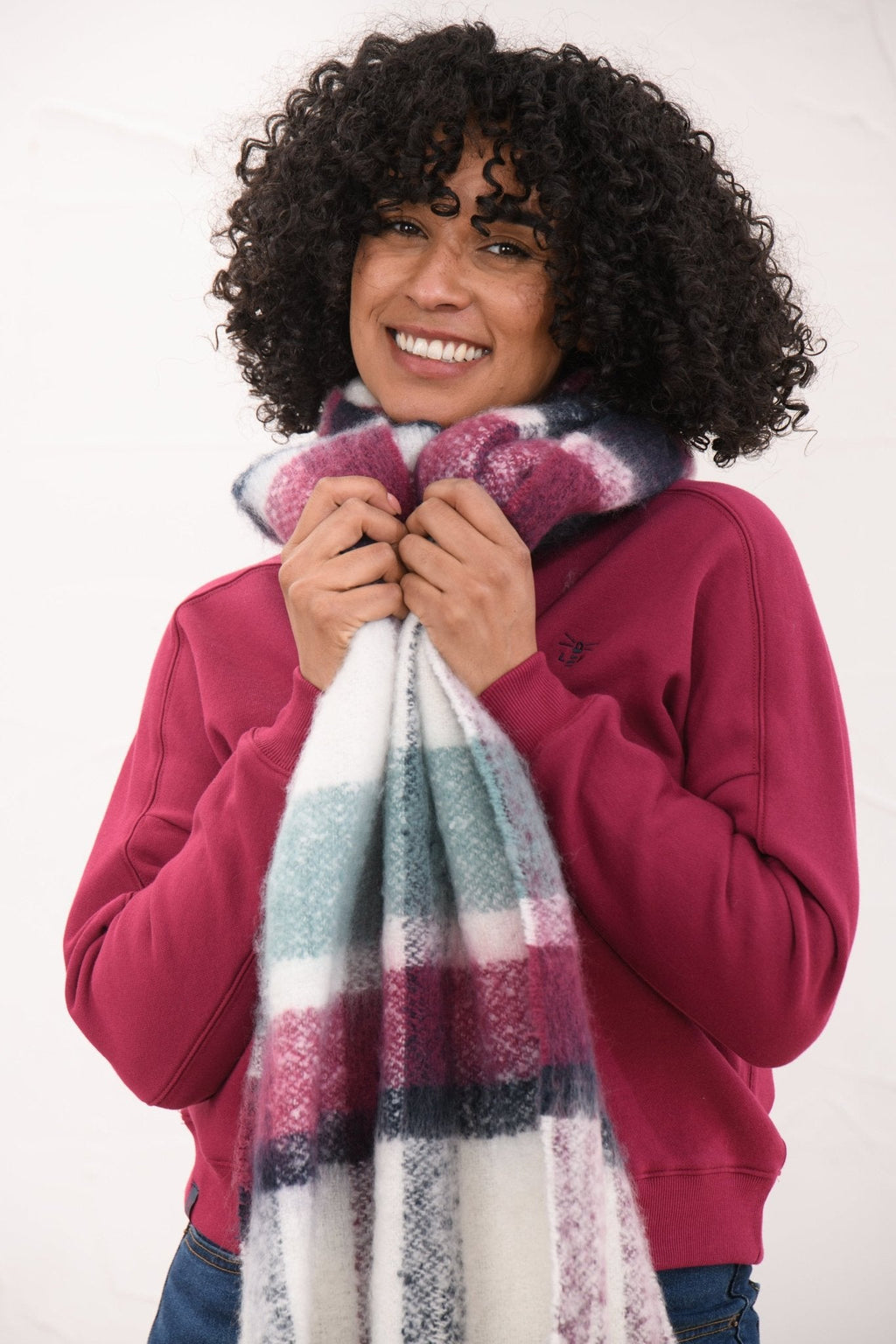 A smiling woman with curly black hair models a thick, soft striped scarf in white, purple, teal, and navy, paired with a magenta sweatshirt against a plain white background.