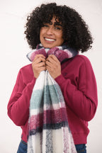 A smiling woman with curly black hair models a thick, soft striped scarf in white, purple, teal, and navy, paired with a magenta sweatshirt against a plain white background.