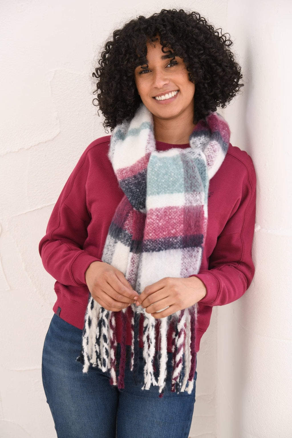 A smiling woman with curly black hair models a thick, multicolor plaid scarf with white fringe. She is wearing a magenta sweatshirt and blue jeans, standing against a white textured wall.
