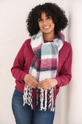 A smiling woman with curly black hair models a thick, multicolor plaid scarf with white fringe. She is wearing a magenta sweatshirt and blue jeans, standing against a white textured wall.