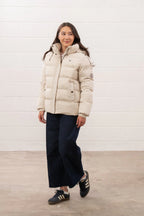 Woman wearing a beige puffer jacket against a white wall.