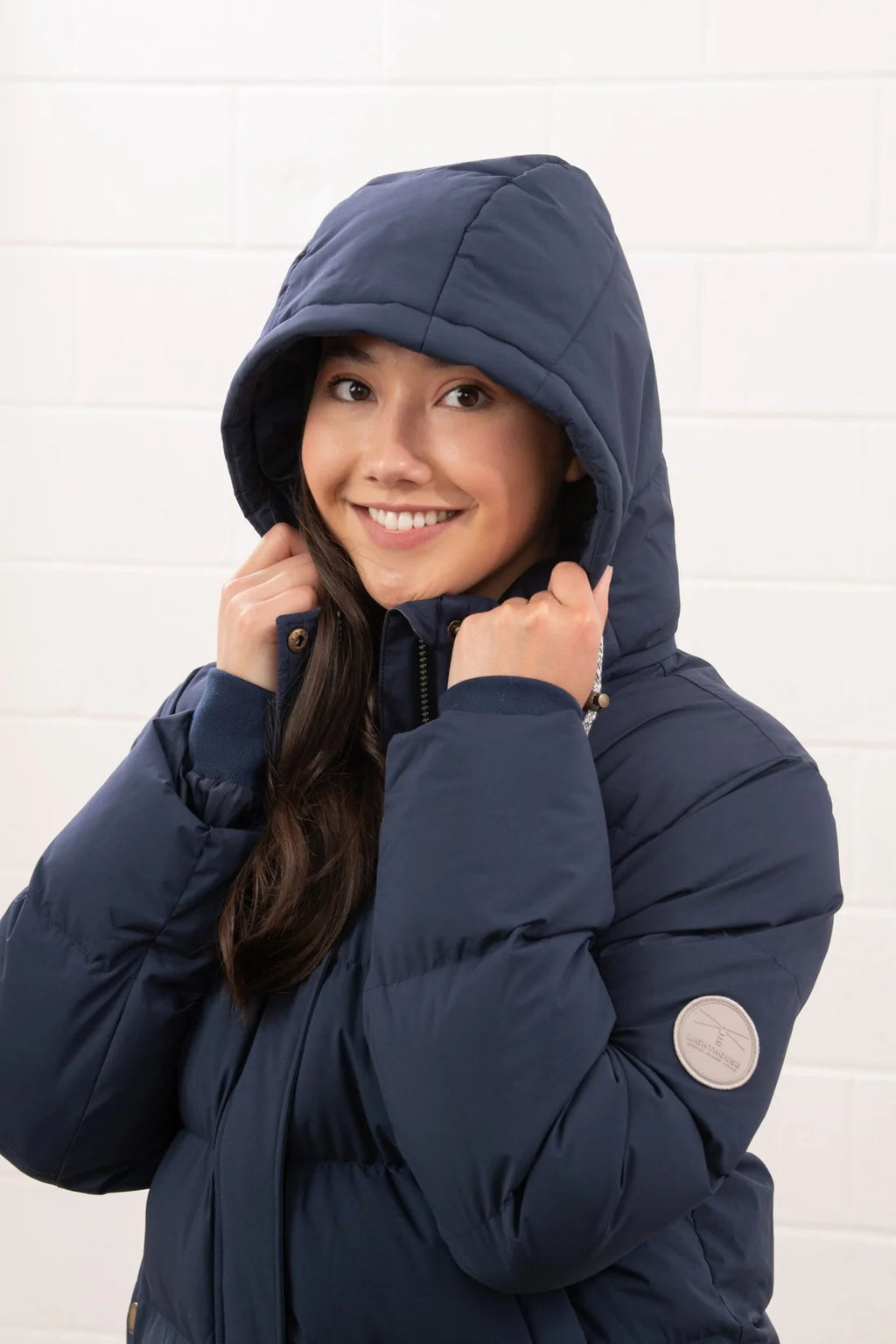 Person wearing a navy blue puffer jacket with a hood against a white brick wall.
