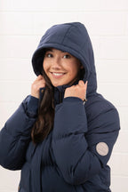 Person wearing a navy blue puffer jacket with a hood against a white brick wall.