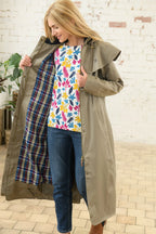 Woman wearing a long green coat over a colorful floral shirt with a plaid shirt underneath, standing in front of a brick wall.