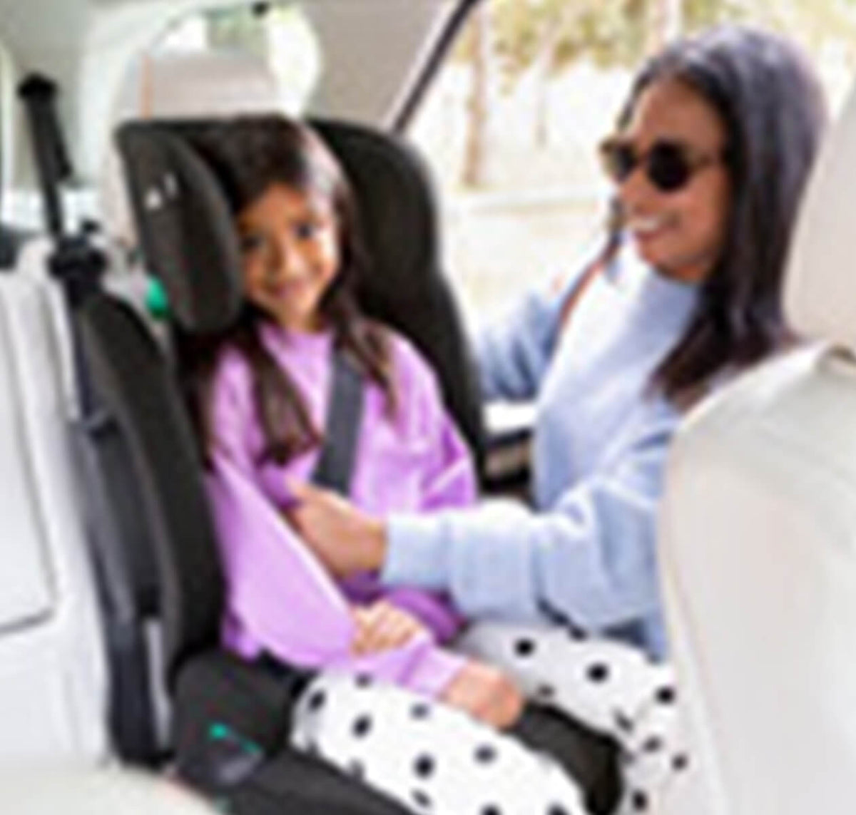 A woman in a light blue sweater and sunglasses is fastening a child's seatbelt in a black car seat installed in a vehicle interior. The child is wearing a purple top and white pants with black polka dots.