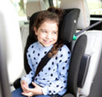 A young girl with brown hair sitting in a black and gray child car seat inside a vehicle, wearing a light blue long-sleeve shirt with black polka dots and a black seatbelt across her chest.