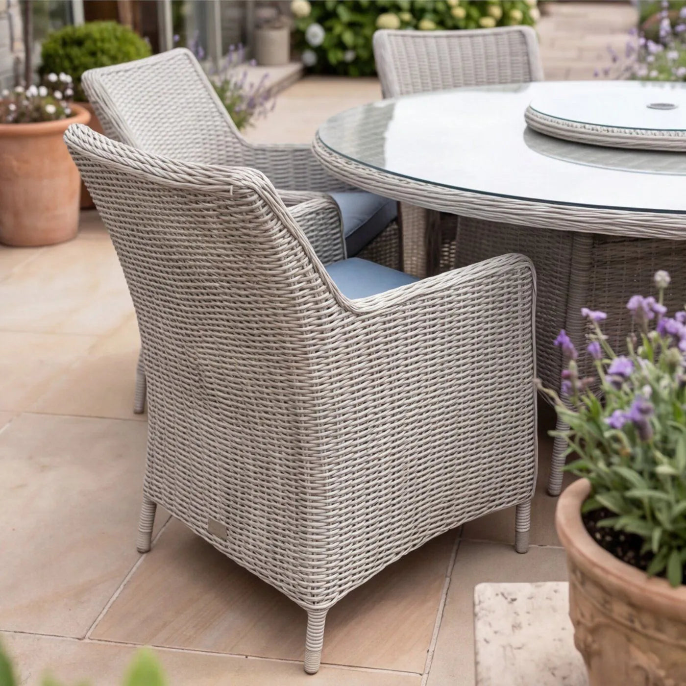 A light gray woven wicker patio dining set with two armchairs and a round glass-top table, shown on a tiled outdoor patio with potted plants.