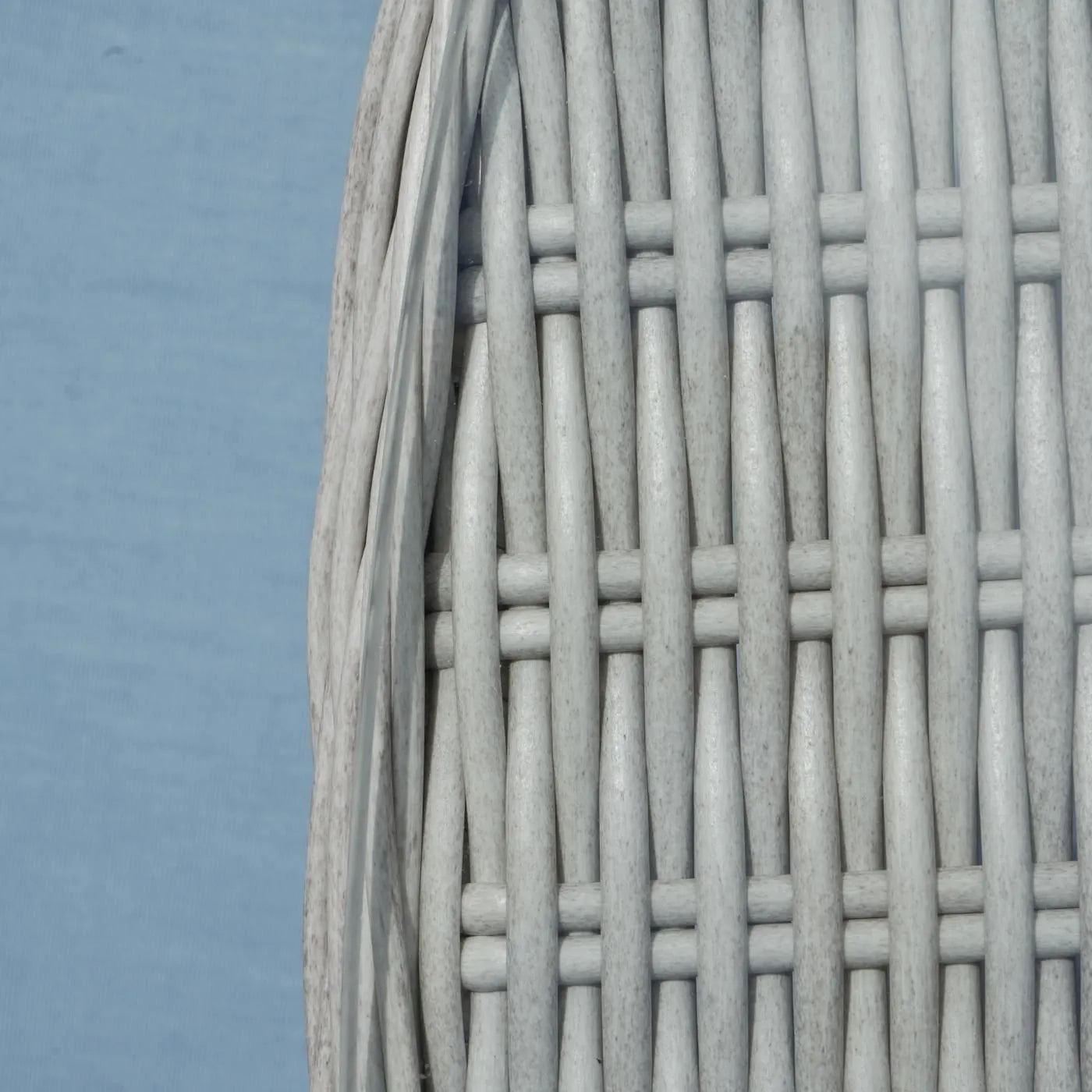 Close-up of a light gray woven wicker or rattan material with vertical strands interlaced horizontally, shown against a pale blue background.