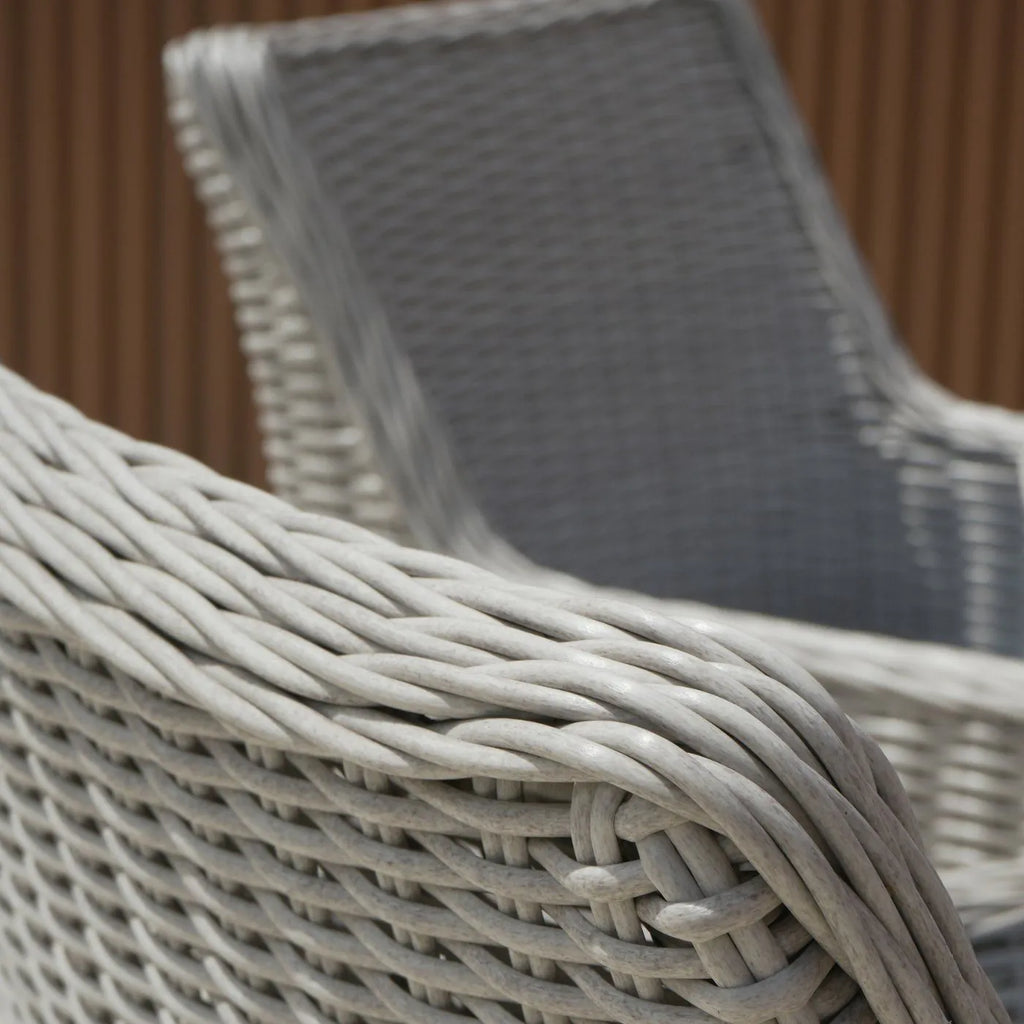 Close-up of a light gray wicker patio chair showing the woven armrest and backrest, with a second matching chair blurred in the background against a brown slatted wall.