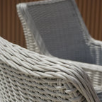 Close-up of a light gray wicker patio chair showing the woven armrest and backrest, with a second matching chair blurred in the background against a brown slatted wall.