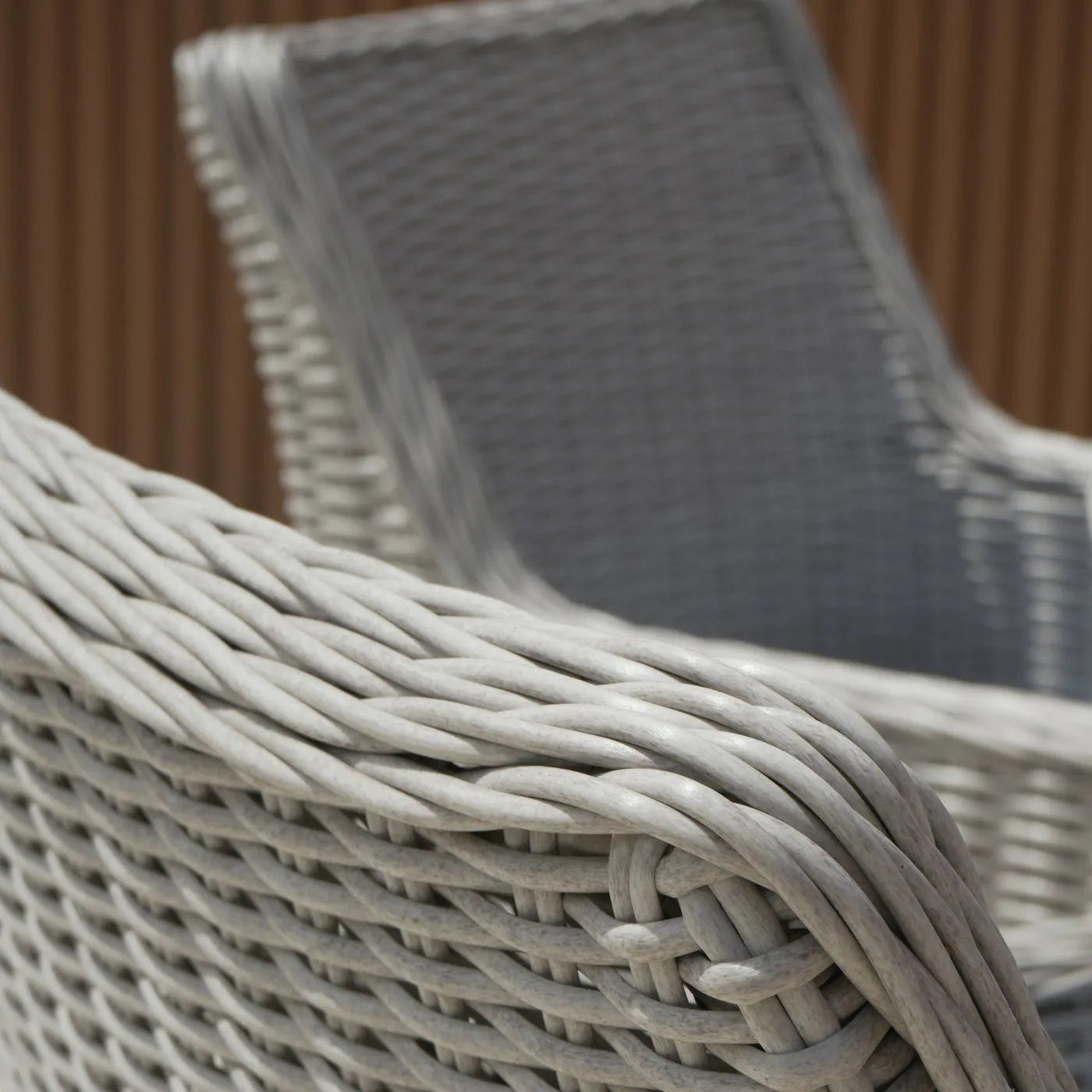 Close-up of a light gray wicker patio chair showing the woven armrest and backrest, with a second matching chair blurred in the background against a brown slatted wall.