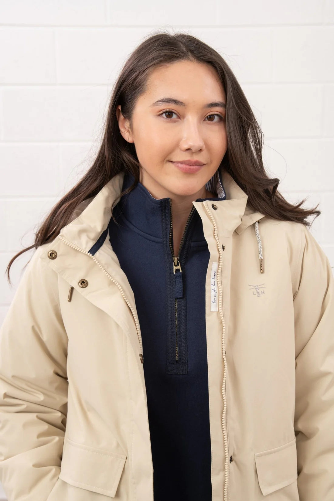 Woman wearing a beige jacket over a dark blue top against a white brick wall.