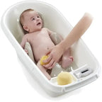 A baby sitting in a white plastic bath tub while an adult hand washes the baby's legs with a yellow sponge. A small digital thermometer is visible on the front right of the tub.